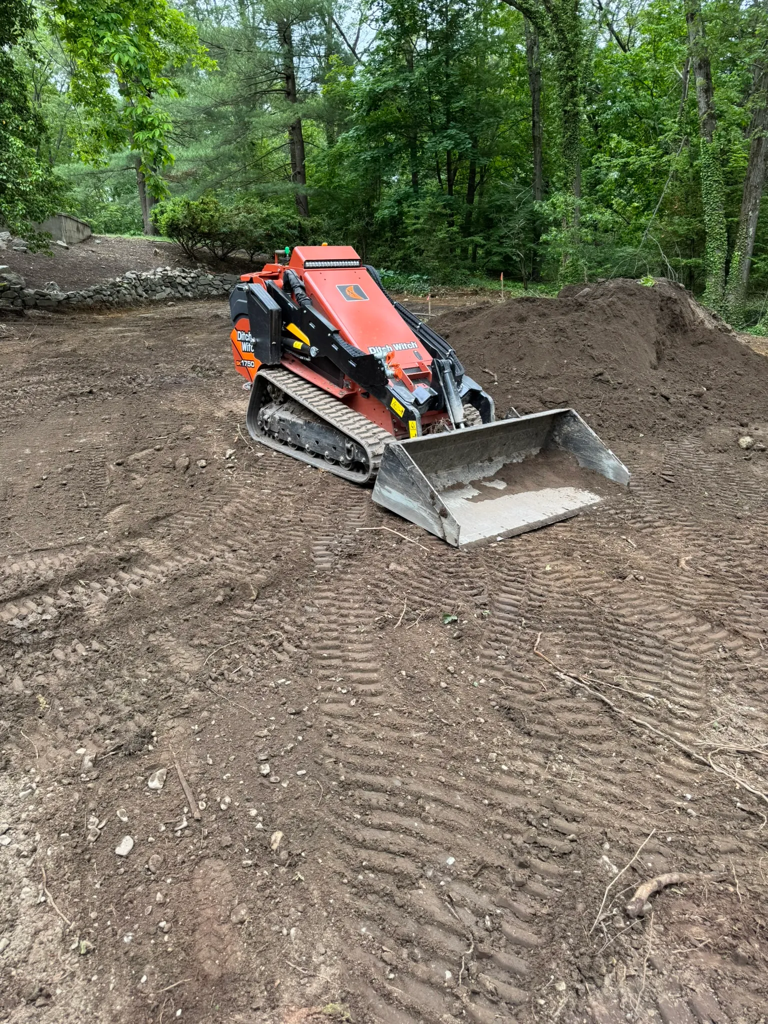A mini skid steer grading a dirt pad beside a fresh pile of soil